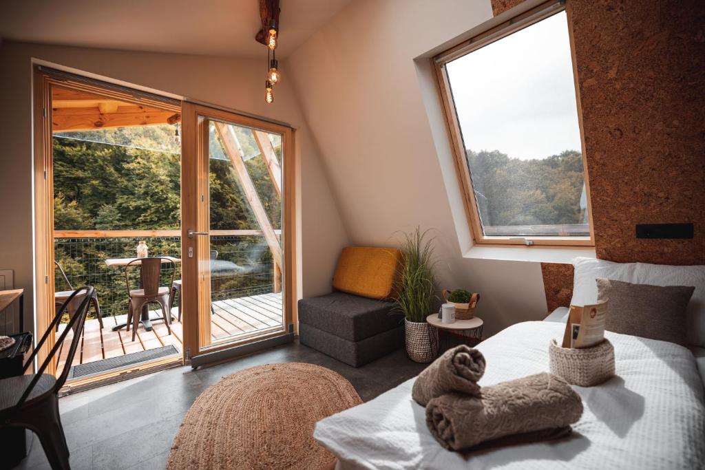 Bedroom with a bed on the right, towels folded on it, and a skylight; glass doors open to a wooden balcony with chairs and trees outside.