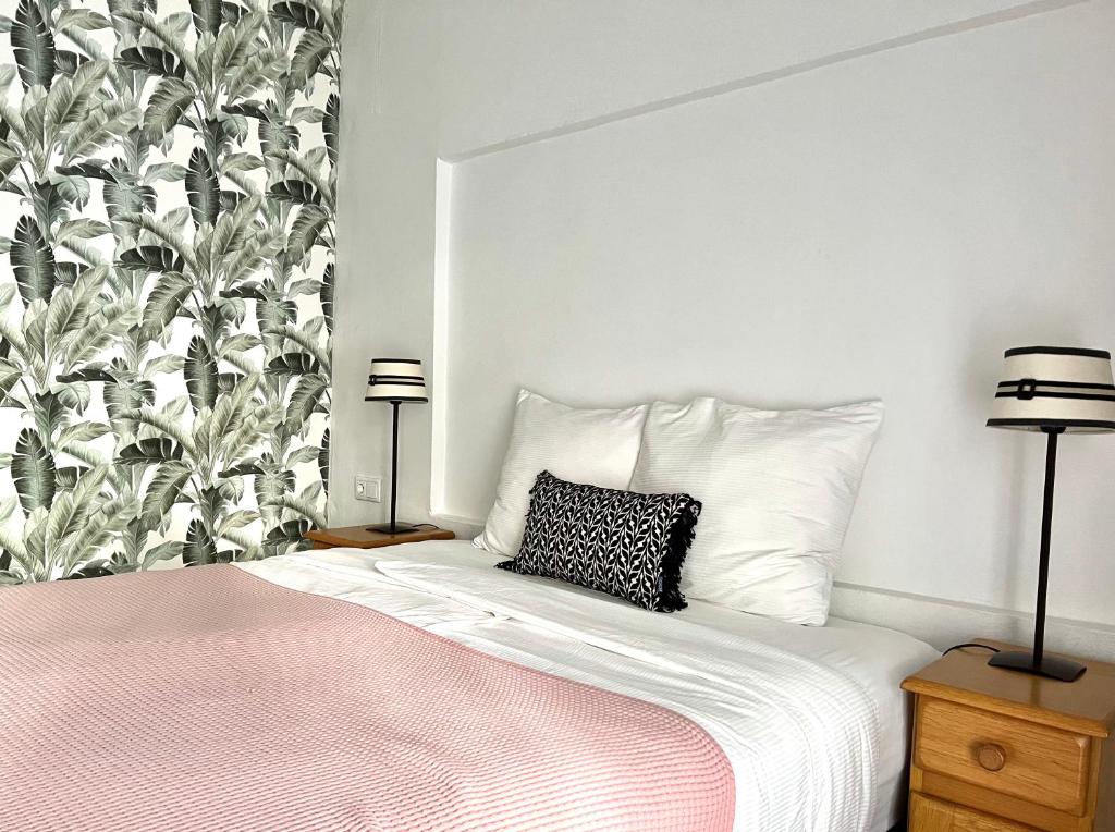 White bed with pink quilt, two wooden nightstands with striped lamps, and tropical leaf wallpaper on the left in a bright, minimal bedroom.