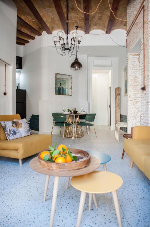 Open living/dining space with mustard sofas, a fruit bowl on a wooden table, terrazzo floor, exposed wooden ceiling beams, a black chandelier, a round glass dining table with green chairs, and a brick wall on the right.