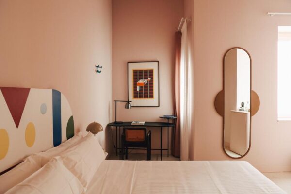 Pastel pink hotel room with white bedding, a colorful headboard, and a black desk with chair beneath wall art.