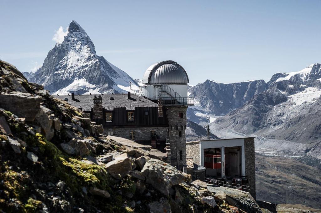 Stone observatory with a silver dome perched on a rocky alpine hillside, snow-capped peaks in the distance.