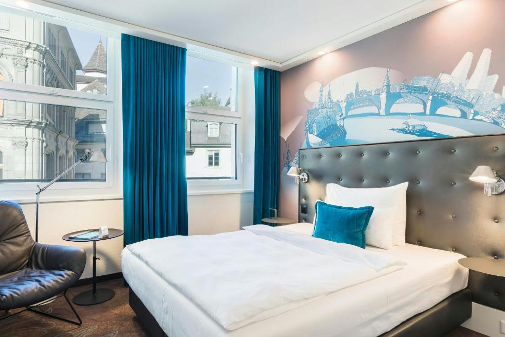 Bright hotel room with a tufted gray headboard, white bedding, and a blue accent pillow, plus blue curtains and a city mural on the wall behind the bed.