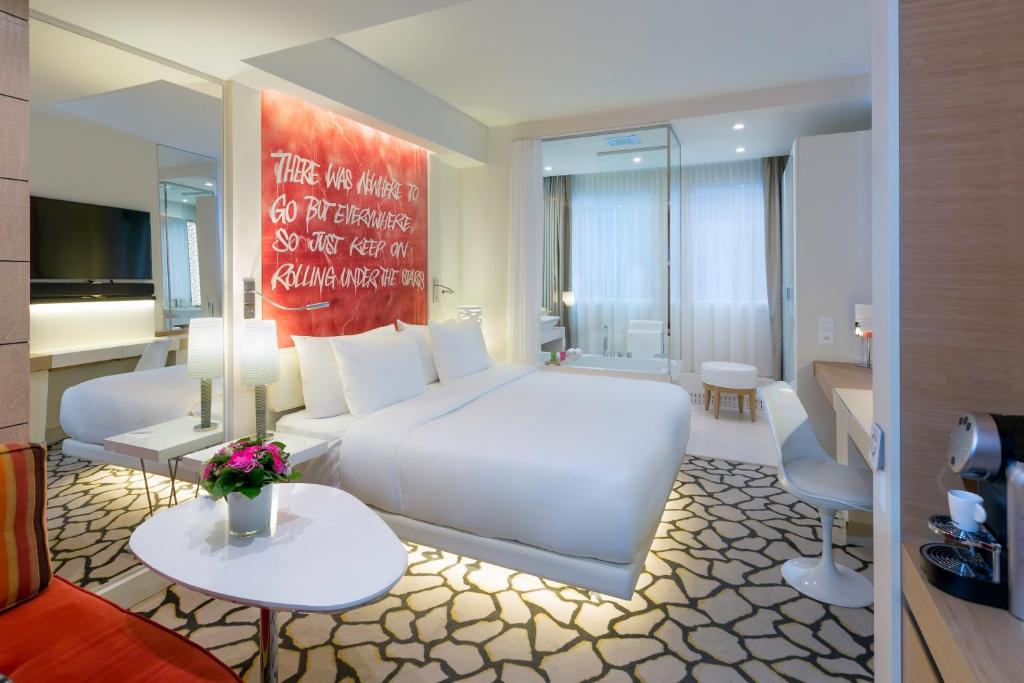 Modern hotel room with a large white bed, red abstract artwork above, glass-enclosed bathroom, and a patterned carpet.