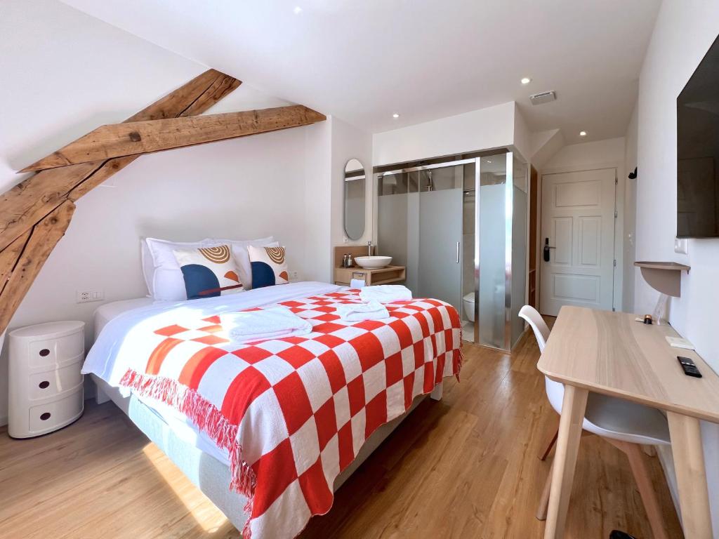 Bright hotel bedroom with an angled wooden beam, a queen bed in white linens and a red-and-white checkered throw, white nightstand, desk, and TV. A glass-enclosed shower is visible in the corner.