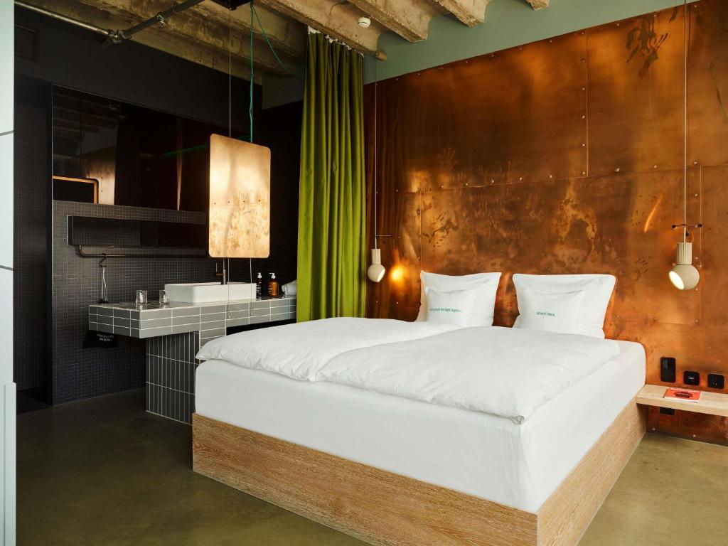 Industrial-style hotel room featuring a king bed with white linens against a copper-toned wall, green curtain and pendant lamps.