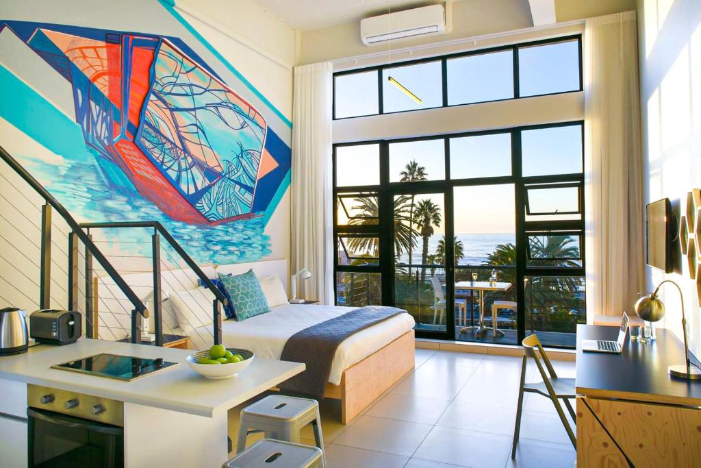 Bright hotel studio with a mural, queen bed, small kitchen, and a balcony overlooking the ocean and palm trees at sunset.