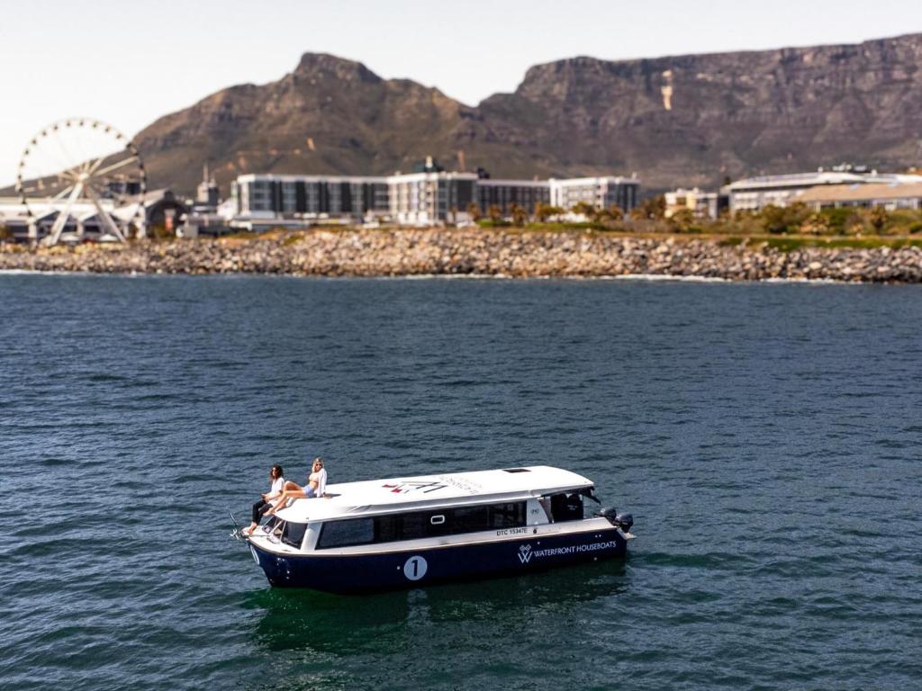 A small white and navy passenger boat with two people on the upper deck, gliding on blue water near a rocky shore and city buildings with a Ferris wheel in the background.