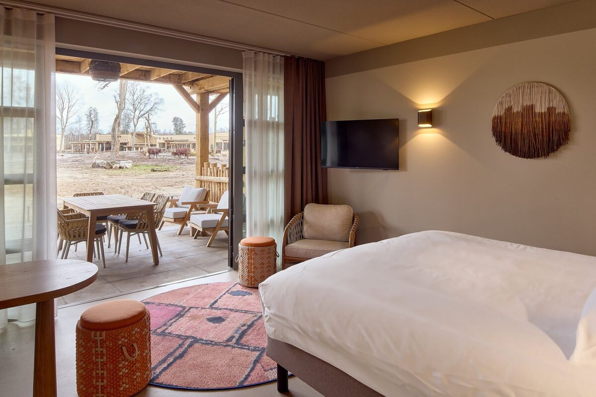 Hotel room with a large white bed and a sliding door to an outdoor patio with a wooden table, chairs, and farm scenery outside.