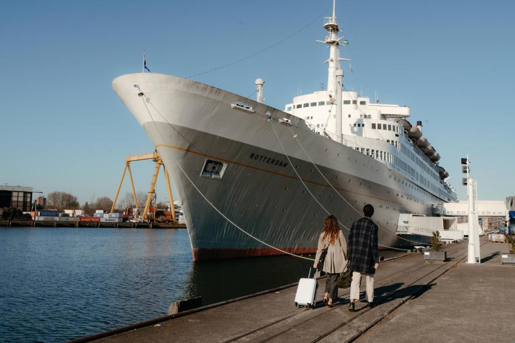 Large white cruise ship moored at a pier, two people with a white suitcase walking toward it.