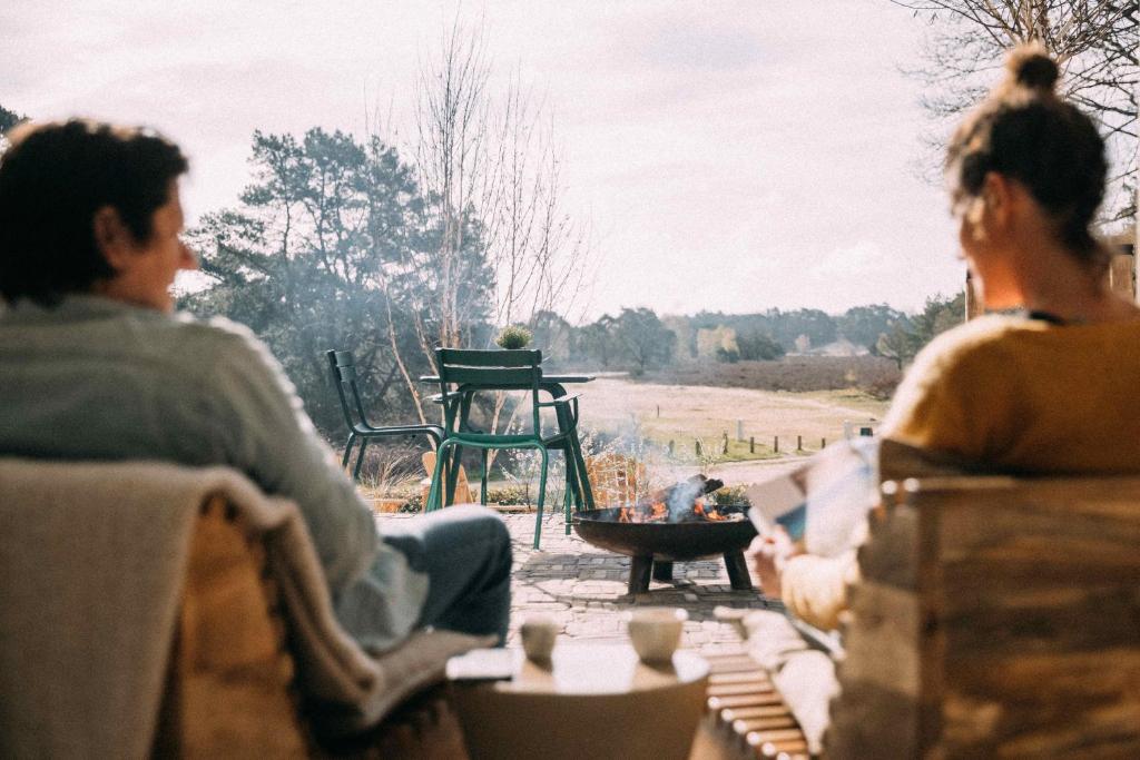 Two people sit around a small fire pit on an outdoor patio, with green chairs and a park in the background.