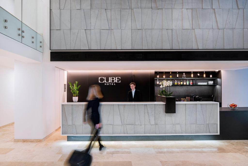 Hotel reception desk with a staff member standing behind it in a modern lobby; sign reads 'CUBE HOTEL' and a guest walks by with a rolling suitcase in the foreground.