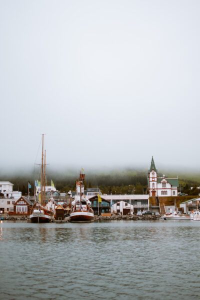 Harbor with sailboats moored at a marina, a white-and-green church on the right, and misty hills in the background.