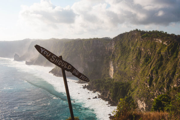 Coastal cliffside with turquoise water and a weathered sign in the foreground that reads 'YOU SHOULD BE HERE'.