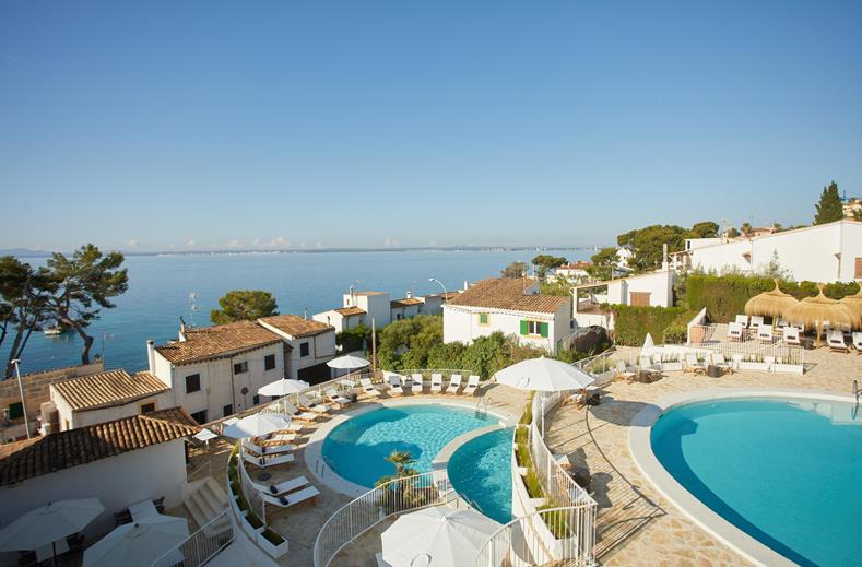 Resort complex with two large pools, whitewashed buildings, and sun loungers overlooking a blue sea.