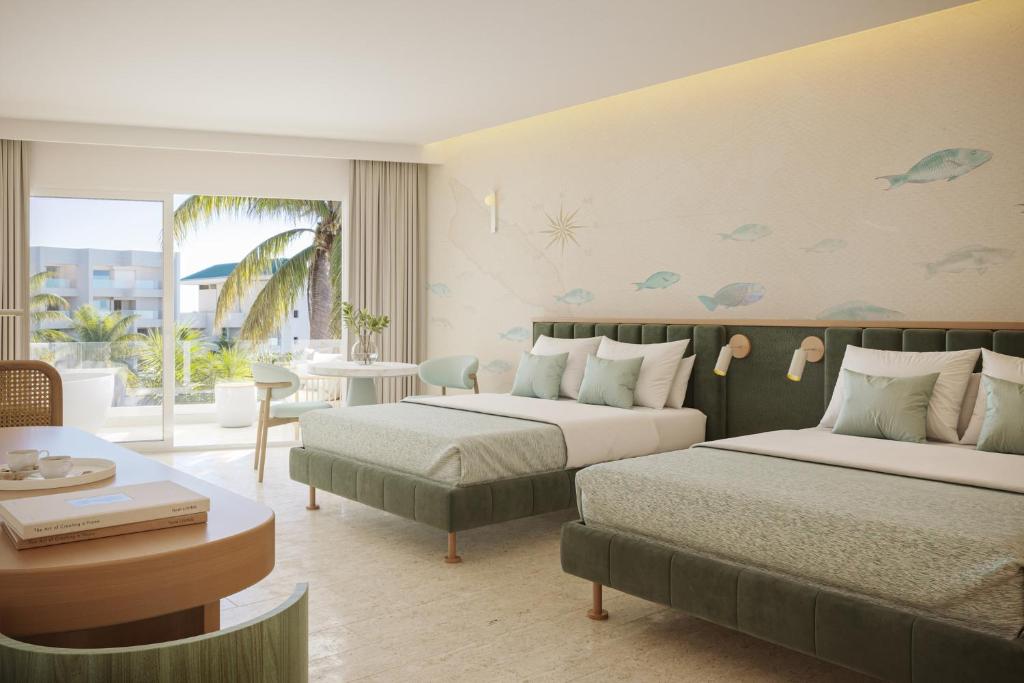 Hotel room with two green upholstered beds, pastel bedding, a sea-themed wall mural, and a balcony with palm trees outside.