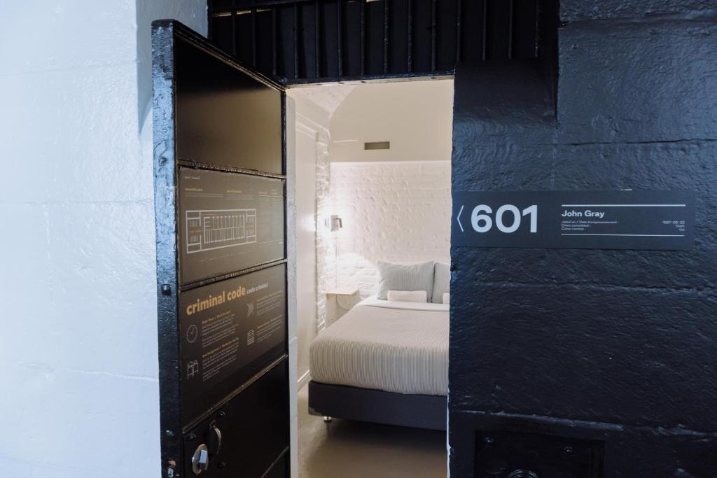 Door open to a white-brick bedroom with a bed, viewed through a dark industrial doorway and sign 601 on the wall.