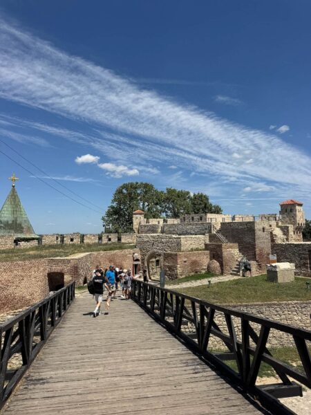 Wooden footbridge leading to a stone fortress on a sunny day, with tourists walking across.