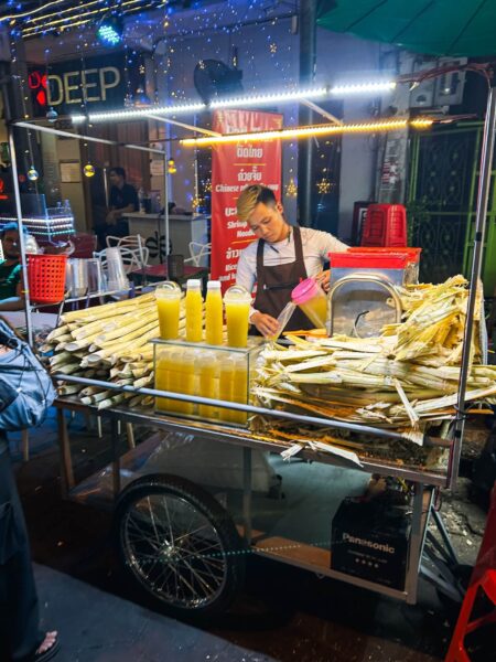 Street food cart at night selling corn on the cob and yellow juice, with a vendor in an apron.