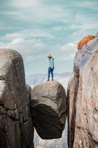 Person in a teal jacket and beige beanie stands on a large boulder wedged between two rocky cliffs, with distant mountains under a blue sky.