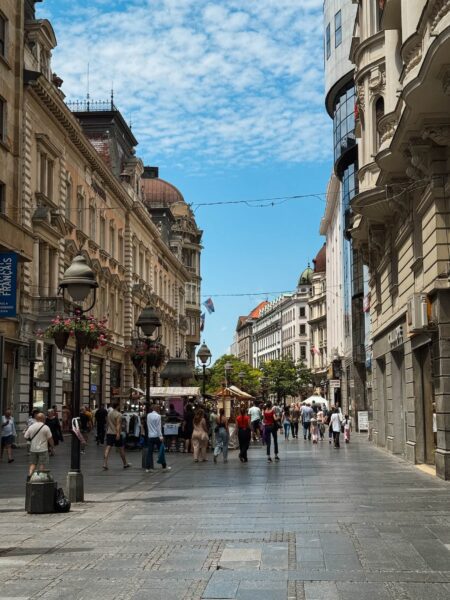 Busy pedestrian street lined with ornate historic buildings and hanging flower baskets, filled with people shopping under a bright blue sky.