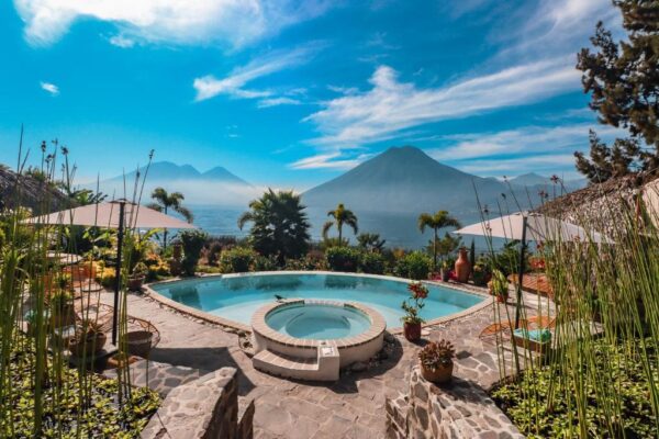 Pool and circular hot tub on a stone terrace with parasols, palm trees, and a sea view with distant mountains under a blue sky