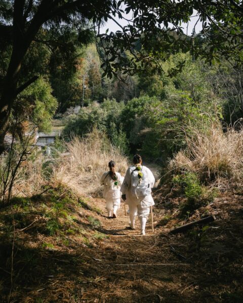 Two people in white outfits walk down a dirt forest path, carrying staffs, with tall grasses and trees on both sides (rear view).