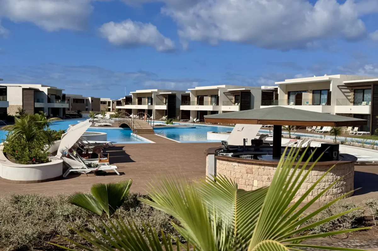 Resort pool area with lounge chairs, white buildings, and palm plants around a central blue pool under a bright sky.