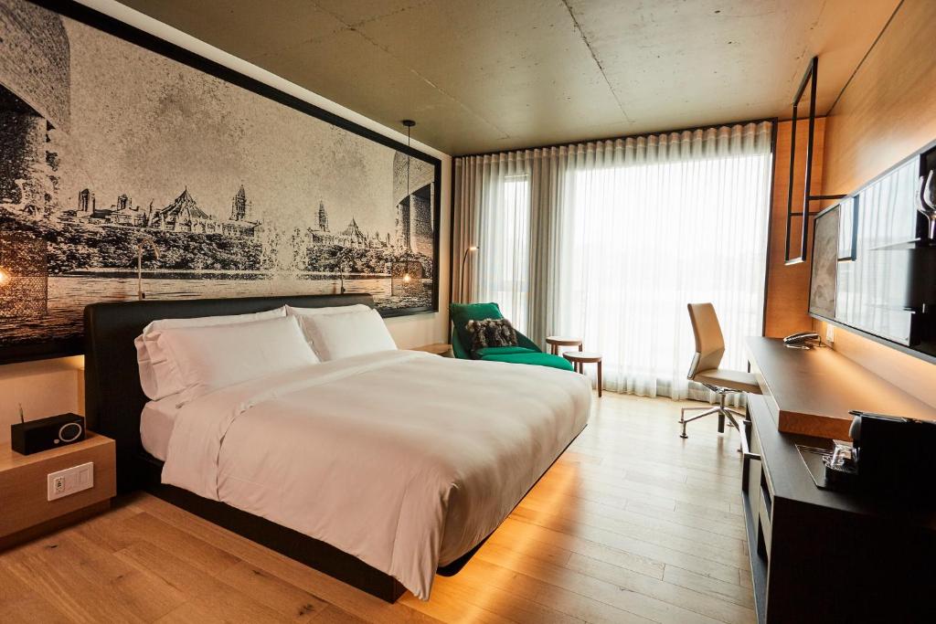 Modern hotel room with a king bed, white linens, a green lounge chair, and a large window with sheer curtains.