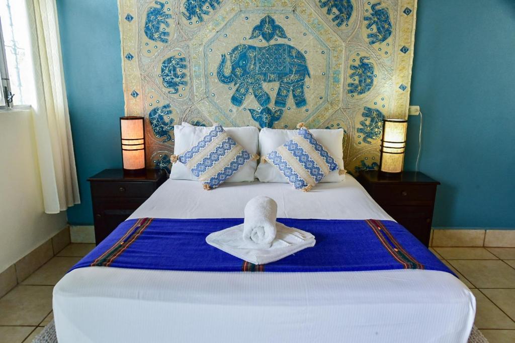 Queen bed with white linens, blue runner, and a rolled towel on top in a turquoise-walled room with an elephant tapestry centerpiece and two bedside lamps.