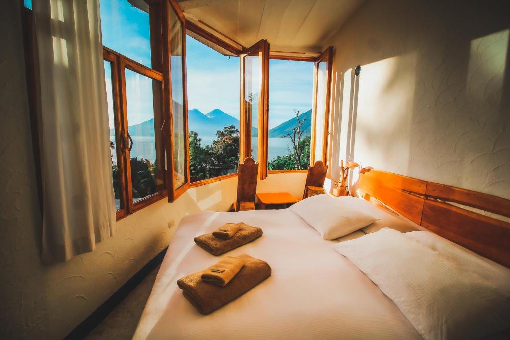 Sunlit hotel room with a large bay window open to a view of mountains and a lake; a double bed is neatly made with white linens and folded brown towels at the foot.