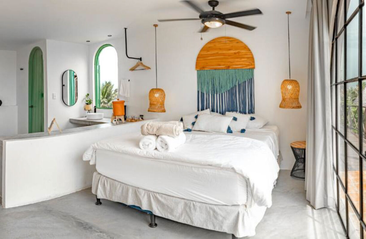 Bright hotel bedroom with white bedding, a green arched door, round mirror, and a large orange-and-teal wall hanging above the bed of the room's wall art.