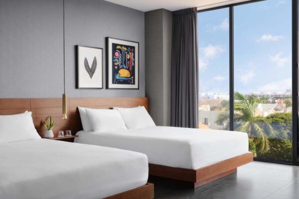Hotel room with two white beds, wooden headboards, framed art, and a large window showing palm trees and a cityscape at dusk.