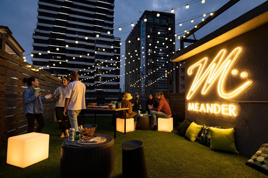 Urban rooftop party at dusk with string lights, people chatting near a neon Meander sign on the wall.