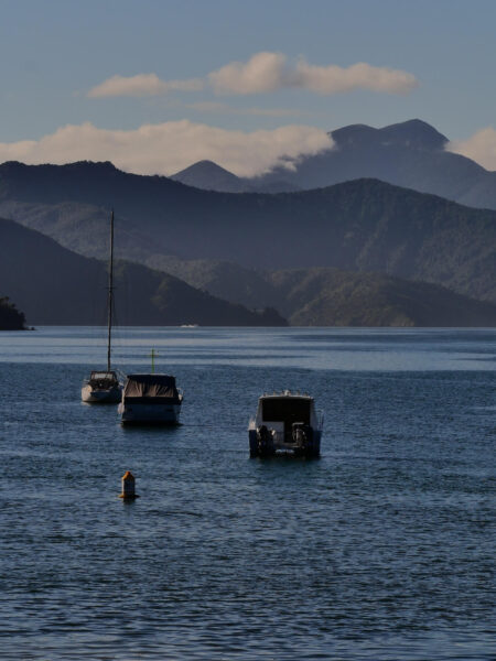 Boats anchored on calm sea with forested mountains and blue sky in the background.
