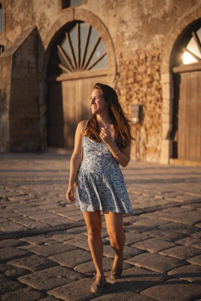 Woman in a blue patterned sundress strolls along a cobblestone path with rustic stone arches in the background, sunlit.
