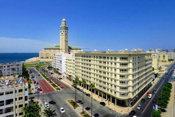A seaside city street with palm trees, a tall minaret-like tower, and a curved beige building along the boulevard.