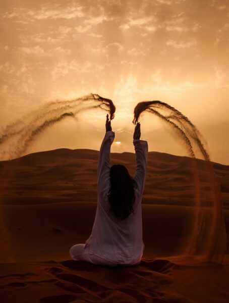 Person sitting in a desert at sunset with arms raised, creating curved trails of sand in the air.