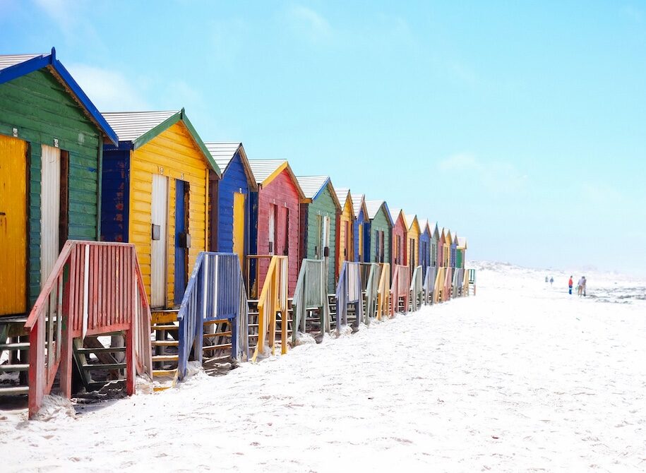 Row of colorful beach huts with stairs leading to doors along a sandy beach on a clear day.