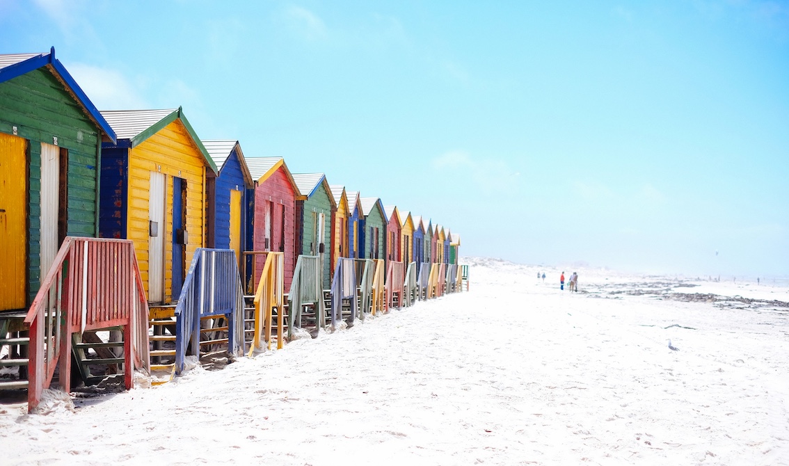 Row of colorful beach huts with stairs leading to doors along a sandy beach on a clear day.