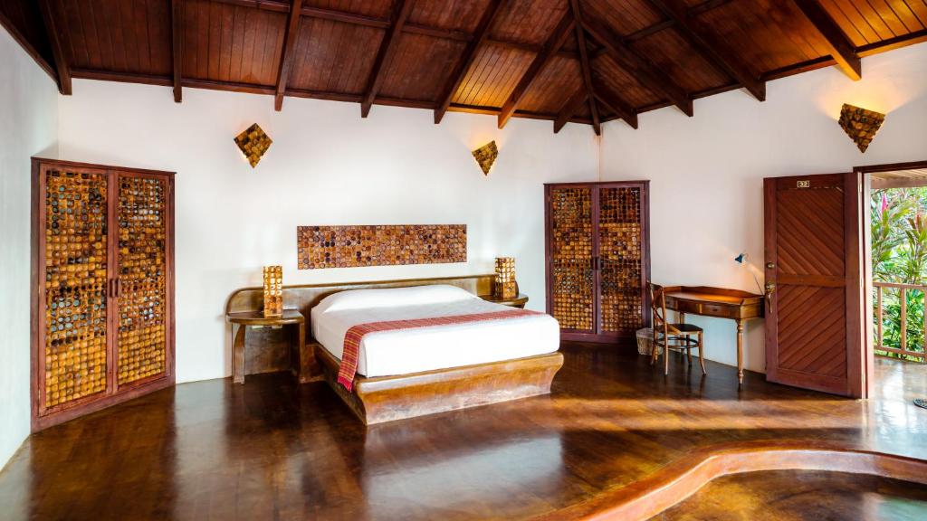 Rustic tropical bedroom with a king bed, white linens, and red accent throw; dark wood floors, vaulted wooden ceiling, and antique wooden wardrobes.
