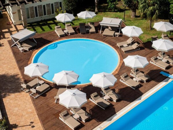 Aerial view of a circular blue pool surrounded by beige lounge chairs and white umbrellas on a wooden deck near a resort building.