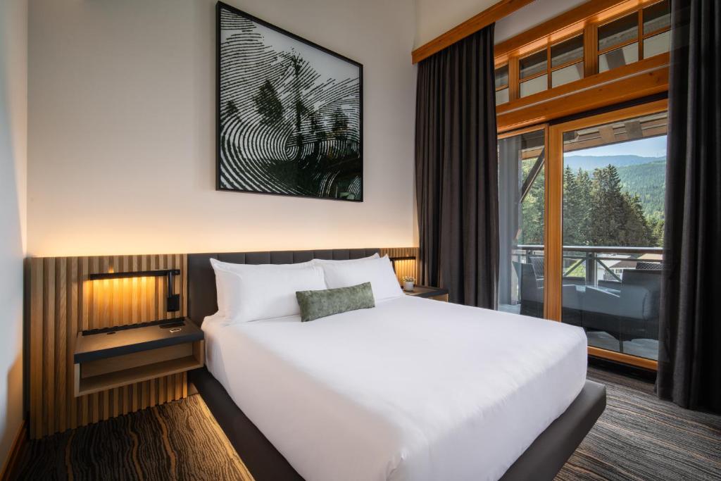 Hotel room with a large white bed, wood headboard, bedside lamps, and a balcony door showing mountains outside.