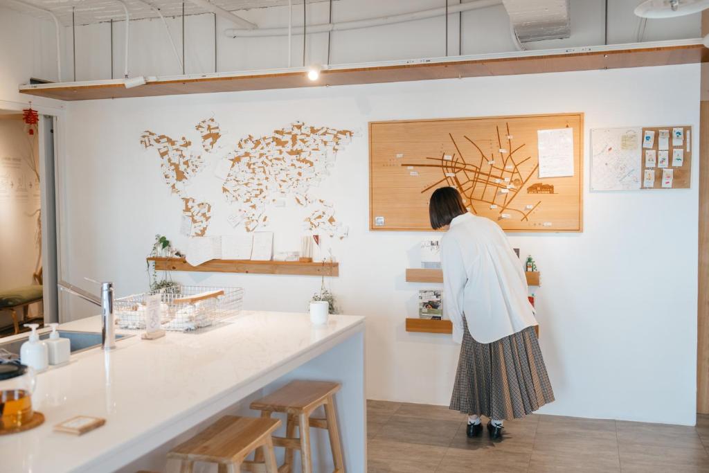 Woman in a white blouse and plaid skirt leans to inspect a large map on a white wall in a bright cafe-like space, with a counter and stools in the foreground.
