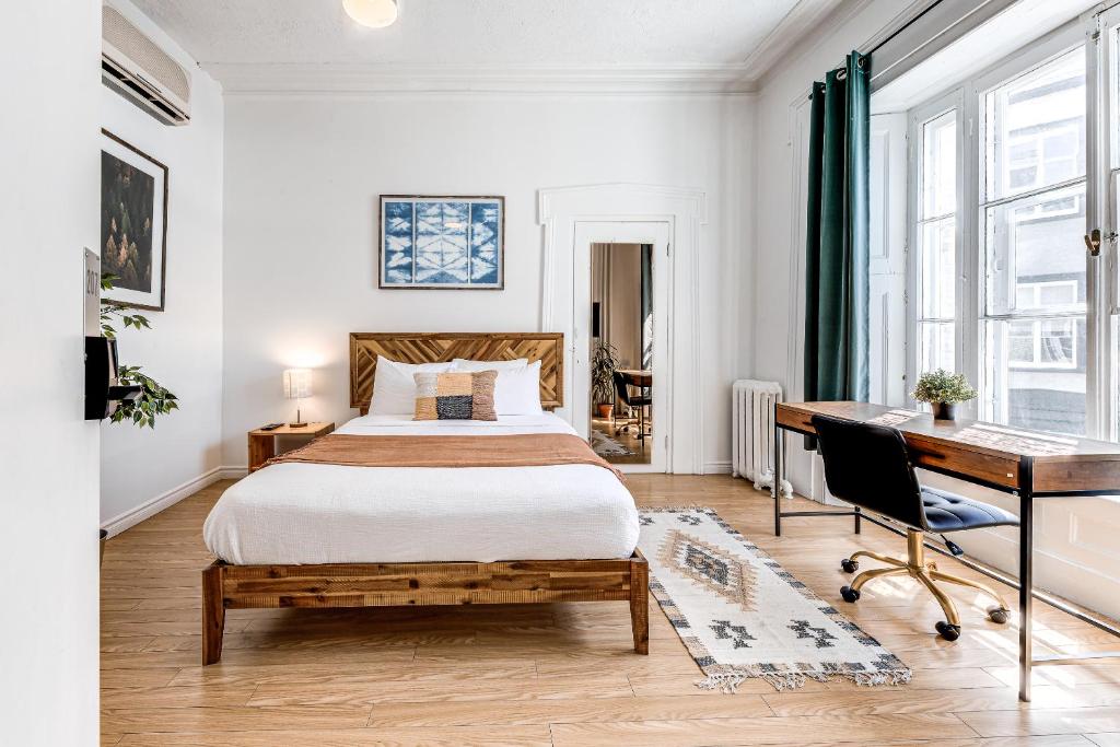 Bright bedroom with a wooden bed, white bedding, and a desk by large windows to the right.