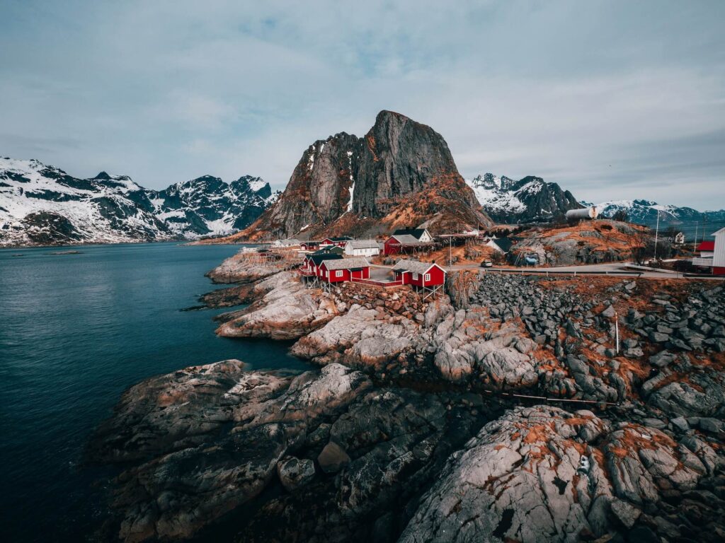 Coastal village with red wooden houses at the base of a tall rocky mountain, snow-capped peaks in the distance and calm blue water nearby to the left.