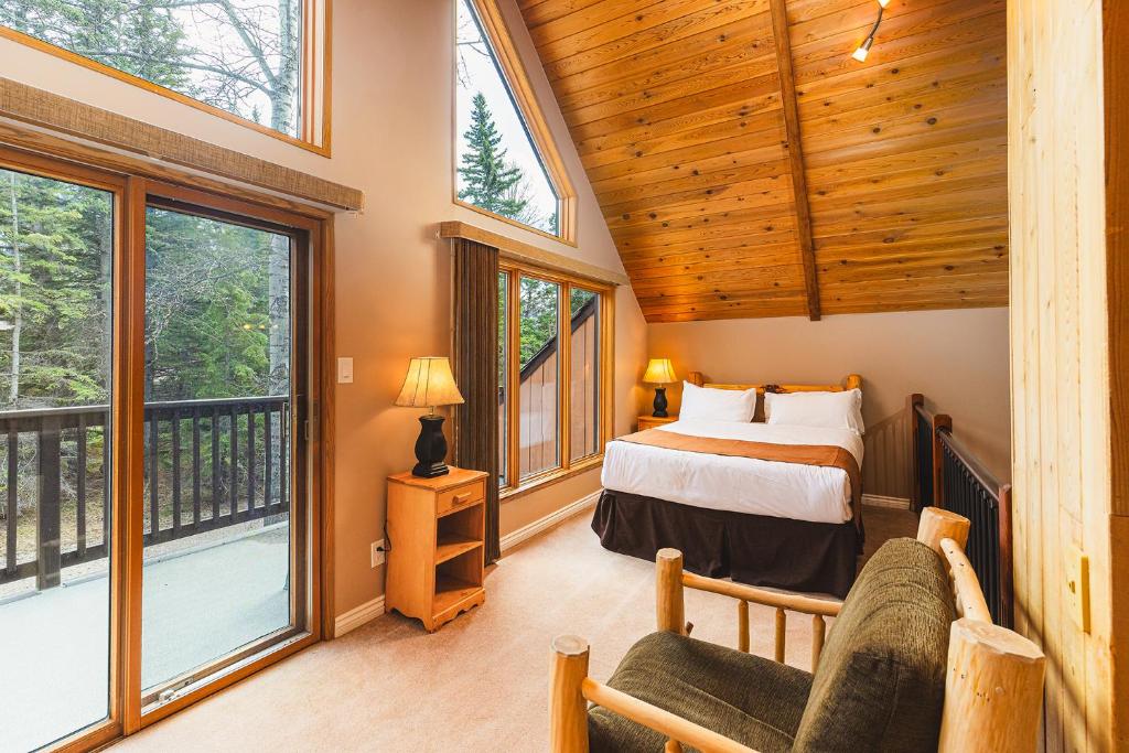 Cozy cabin bedroom with a vaulted wooden ceiling, large windows and a balcony door, a king bed with white linens and brown accents, and a green armchair on the side.