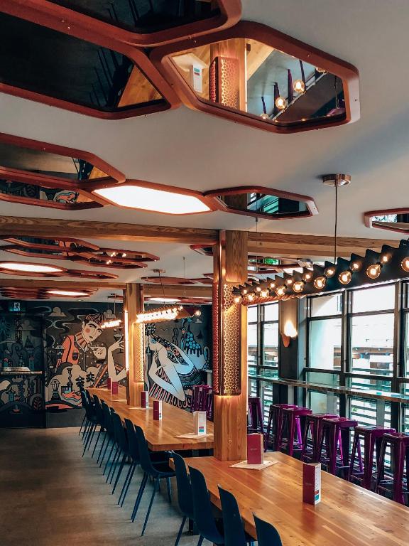 Long communal wooden table with dark chairs in a modern cafe, mural wall, and round pendant lights by tall windows.