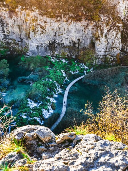 A narrow wooden boardwalk winds through a turquoise river canyon with rocky cliffs and lush green vegetation; a person walks along the path.