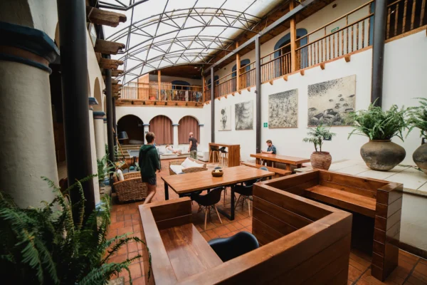 Indoor atrium cafe with wooden tables and benches, potted ferns, and arched doorways on a brick floor under a glass-panel roof.