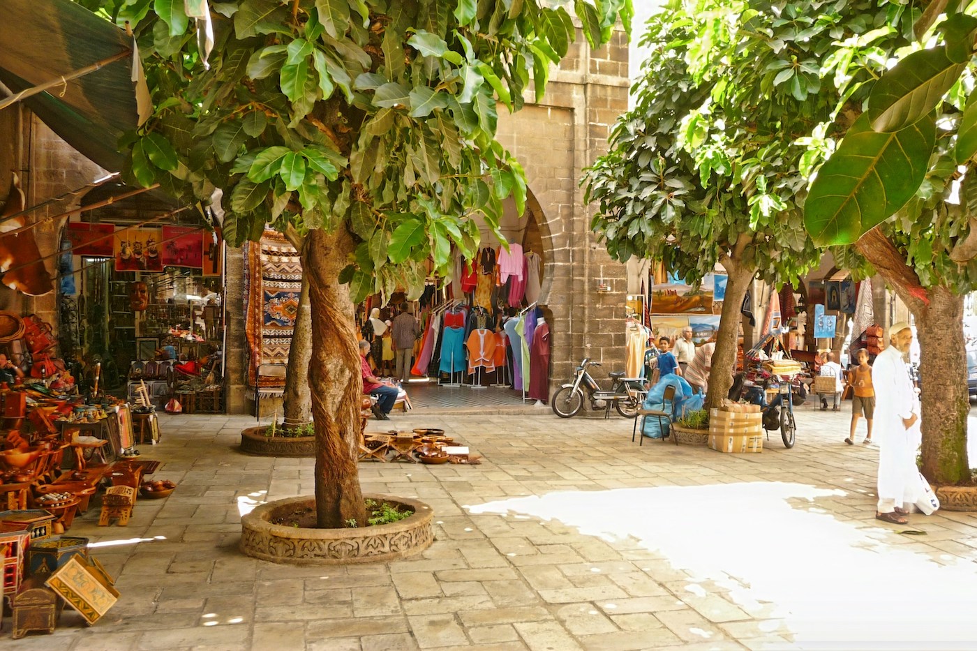 Open-air market under large trees along a stone arcade; colorful clothing, rugs, and crafts for sale.